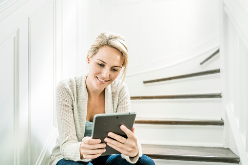 A photo of happy woman using table computer at home. Young female is surfing on digital tablet. Attractive lady is sitting on steps. She is wearing casuals.