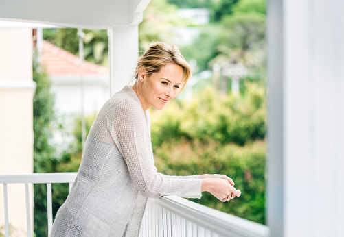 A side view photo of happy woman standing on balcony. Thoughtful young female is in casuals at home. Attractive lady is looking away. She is standing at brightly lit area.