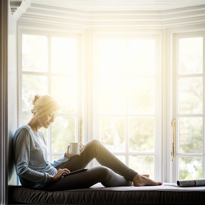 A full length photo of woman using digital tablet at home. Young female is holding coffee cup while sitting on window sill. She is wearing casuals in brightly lit room.