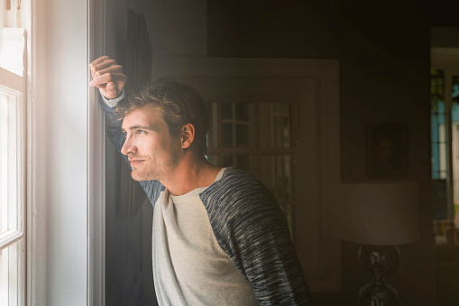 A side view photo of young man looking through window. Thoughtful male standing by window. He is wearing casuals in brightly lit area.