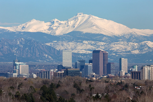 Snow covered Longs Peak, part of the Rocky Mountains stands tall in the background with the Downtown Denver skyscrapers as well as hotels, office buildings and apartment buildings filling the skyline.