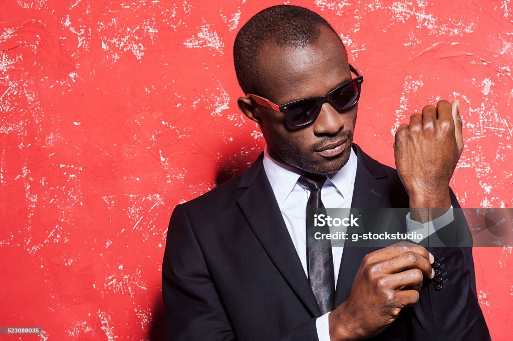Everything should be perfect. Serious young African man in formalwear and sunglasses adjusting his sleeve while standing against red background Fashion Model Stock Photo