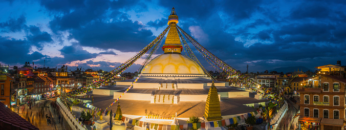 The iconic mandala dome of Boudhanath stupa, illuminated by spotlights under colourful prayer flags fluttering in the evening breeze as crowds of pilgrims and tourists walk around the ancient Buddhist shrine, a UNESCO World Heritage Site in Kathmandu, Nepal's vibrant capital city. ProPhoto RGB profile for maximum color fidelity and gamut.
