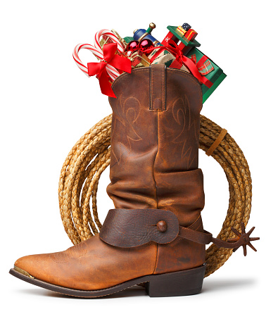 A cowboy boot with a spur attached is stuffed with Christmas presents, a toy train, and candy canes on white background. There is a lasso behind cowboy boot. Clipping path included.