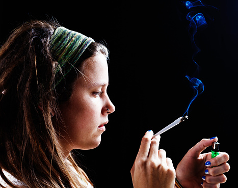 Profile of a pretty young woman with pierced nose and dreadlocks holding a marijuana cigarette, lighter in her other hand.  Black background with copy space.