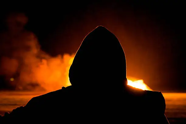 Silhouette of Arsonist in a hooded top looking at a large fire.