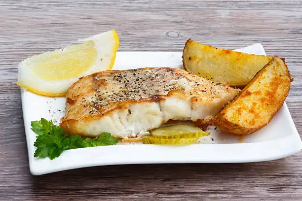 White fish with potato wedges, lemon and celery on white plat, wooden background