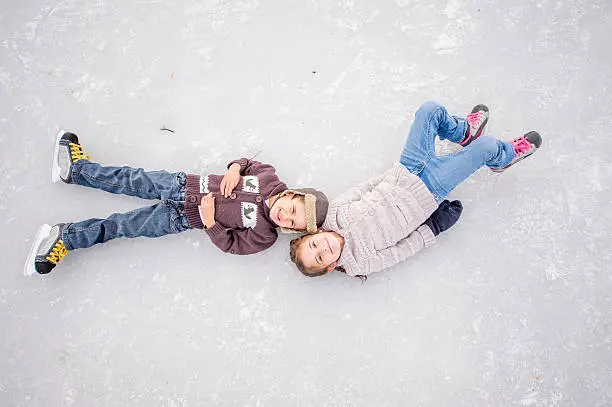 A brother and sister are lying on a frozen lake and are looking up at the camera. The children are wearing ice skates and sweaters.