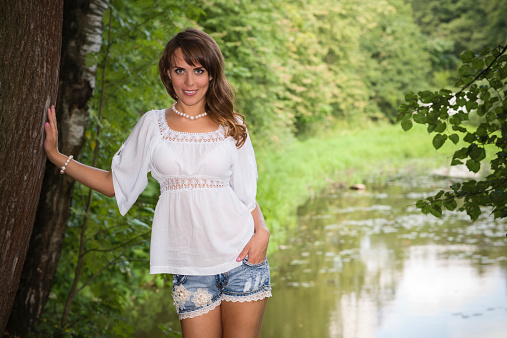 Smiling long haired young woman in cutoffs and white tunic, leaning her arm on a tree and looking at camera, pond in background