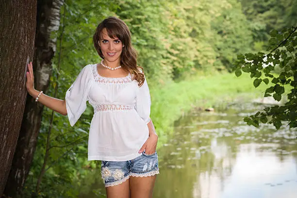Smiling long haired young woman in cutoffs and white tunic, leaning her arm on a tree and looking at camera, pond in background