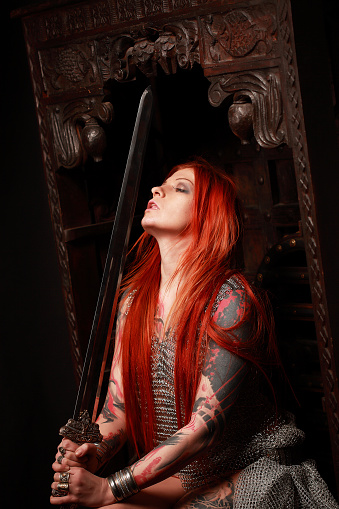 Studio portrait of young attractive redhead girl with lots of tattoo, dressed in chained armour with fantasy sword in her hand