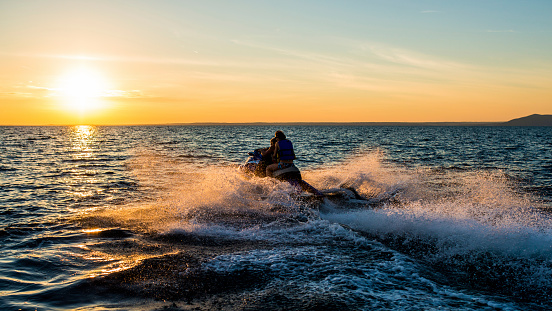 People having fun riding a person watercraft/water scooter on the sea in the sunset.  