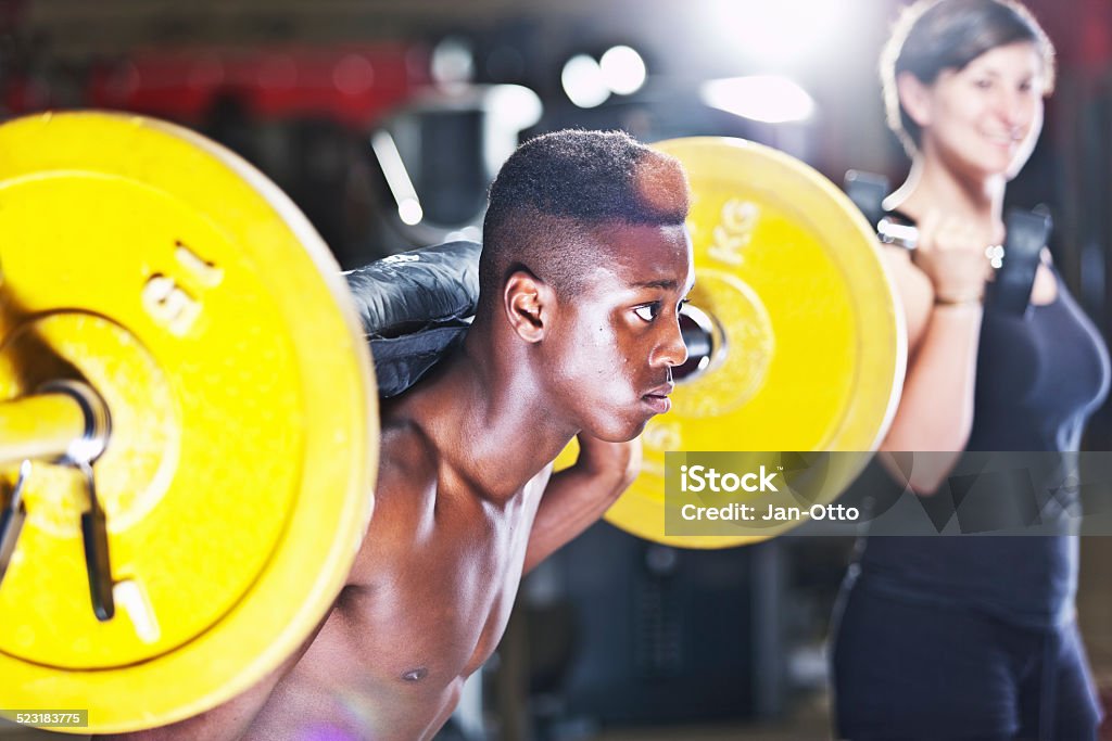 Black musculous guy and caucasian woman in fitness studio A musculous young guy with barbells and a caucasian woman with dumbbelles in a fitness studio. XXL size image. 20-29 Years Stock Photo