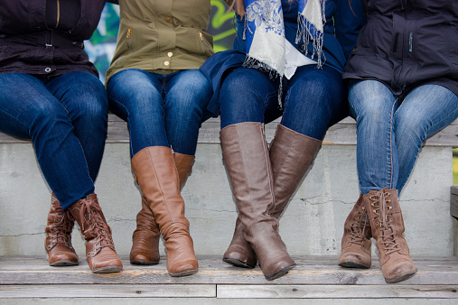 Four pairs of young women's legs crossed at ankle, wearing jeans and boots, sitting outdoors on bleachers