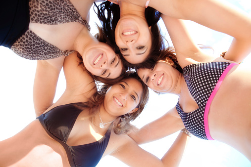 Subject: Horizontal view from below of four pretty Latina young women, hugging close together in a huddle and looking down at the camera. They wear bikinis and swimsuit swimwear as they smile for the camera.