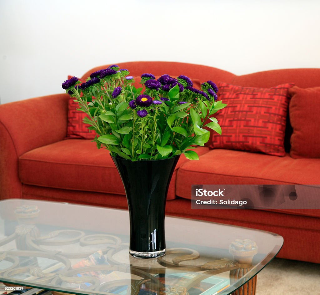 Just A Vase Of Purple Flowers And An Orange Sofa Comfortable contemporary orange living room sofa with cushions in background. Vase of purple Chrysanthemums  in tall black vase on glass table. Blank white wall. Black Color Stock Photo