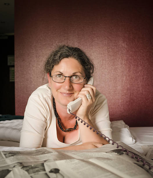 woman laying on bed talking on telephone and reading newspaper stock photo