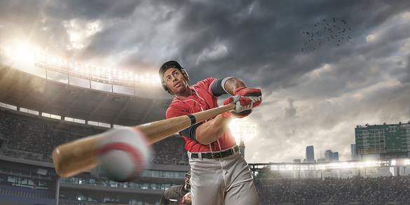 Action extreme close up image of a professional baseball player batting ball during a baseball game. Batter is standing in generic floodlit outdoor baseball stadium full of spectators under stormy evening sky at sunset. Batter is wearing unbranded generic baseball kit and safety helmet. With intentional lensflare and motion blur. 