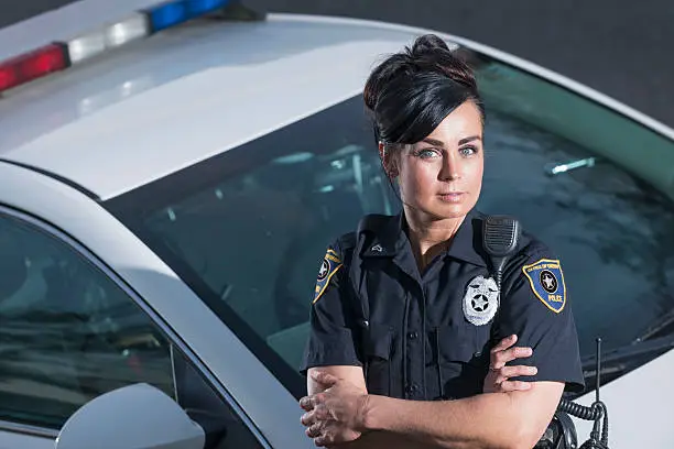 A female police office standing next to her patrol car with her arms crossed, looking at the camera with a serious, confident expression.