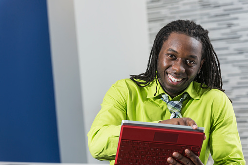 A young, African American entrepreneur, holding a digital tablet, smiling and looking at the camera.  He has dreadlocks and is wearing a bright lime-green shirt and tie.