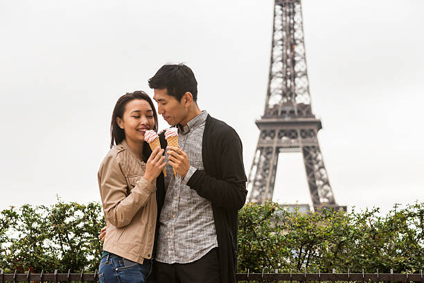 Romantic Couple, a Man with black hair tied in a braid, wearing a blue jacket, a woman with blond hair, wearing a black jacket, a white hat and skirt, sitting on a concrete wall at seine river in front of Eiffel Tower, Paris with lighting in the evening, boyfriend hugging his girlfriend, Woman leans her head against the shoulder of her boyfriend, rear view, focus on forefront, horizontal