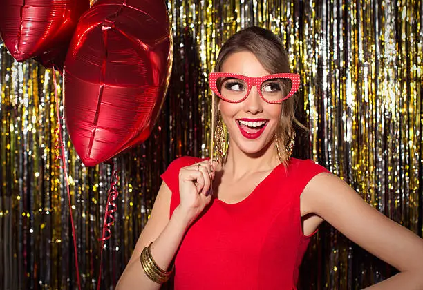 Young woman wearing elegant red dress celebrating or having party in front of fringe curtain. She is holding photo booth fake glasses and posing at camera.