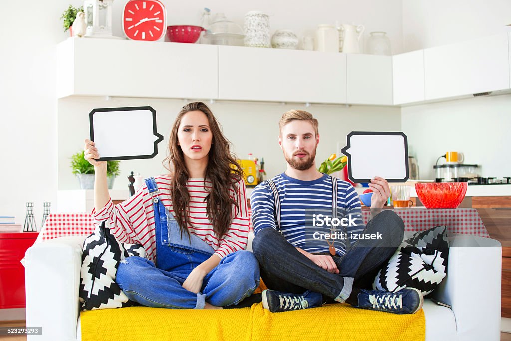 Young couple with speech bubble Young couple in conflict sitting on sofa in their apartment and holding speech bubbles in hands. Kitchen in the background. Couple - Relationship Stock Photo
