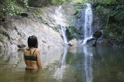 Girl wearing bikini admiring view of beautiful tropical jungle waterfall in Khao Lak national park, Phang Nga, Thailand