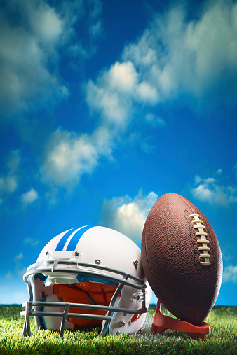 This is a photo of an American football leaning against a white helmet with a sunny cloudscape for copy space in the background.