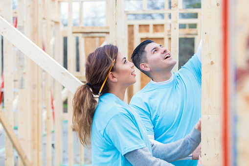 Hispanic couple work together to build charity home. The man shows the woman something on a piece of plywood. They are wearing blue volunteer t-shirts. The woman has a pencil behind her ear. They are working on the frame for the house.