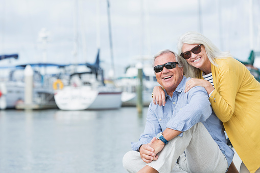 A happy senior couple wearing sunglasses, enjoying a sunny summer day on the waterfront. They are on the boat dock at a marina, smiling and looking at the camera. They are happily retired or on vacation.