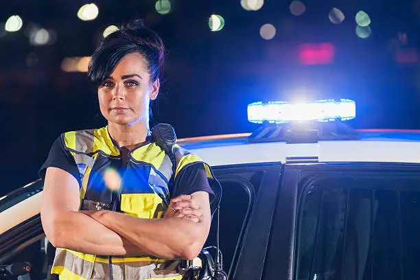 A female police officer wearing a safety vest, standing next to her patrol car, looking at the camera with a serious expression on her face. It is night, and the emergency lights on the police vehicle are flashing.