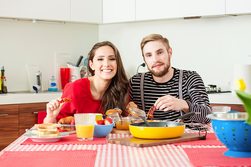 Young couple sitting at the table in the kitchen and eating breakfast, smiling at camera. 