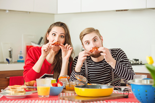Young couple sitting at the table in the kitchen and having breakfast, making bacon mustaches and funny faces.