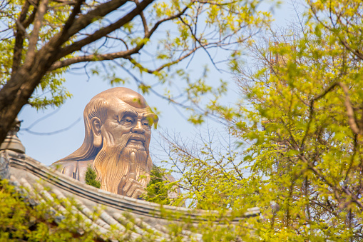Laoshan,China - April 21, 2016:  A huge 36m statue of the spiritual creator of Daoism Lao Tse seen through a forest above a Chinese roof