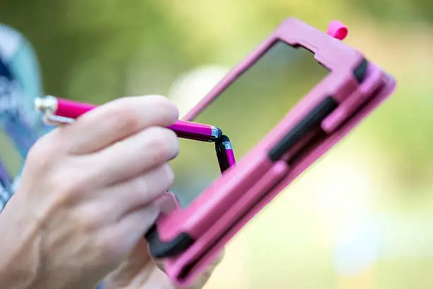 Close up view of the hand of a woman standing outdoors writing on the touch screen of a pink tablet with a stylus.