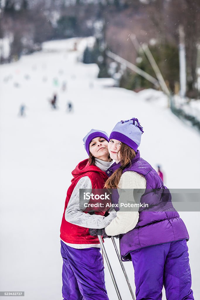 Two sisters posing at the slope at the Ski Resort Two sisters pose together at the bottom of a snowy ski slope at a winter resort as snow falls. Both girls are wearing toboggan hats and brightly colored parkas. One girls is holding two ski poles in her hands. They are posing and smiling in an exaggerated manner looking directly at the camera.  14-15 Years Stock Photo