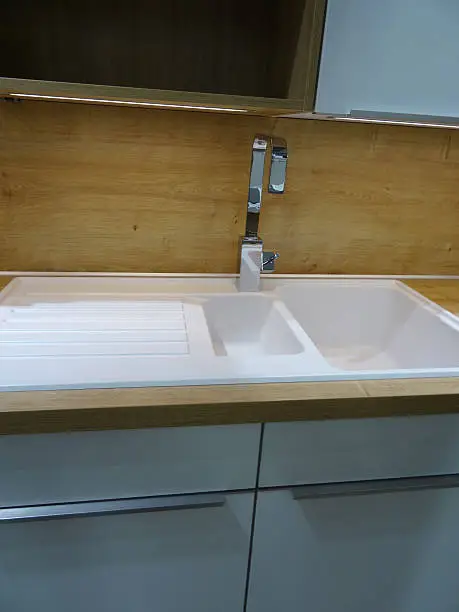 Photo showing a modern double white ceramic kitchen sink and drainer / draining board with a brushed stainless-steel mixer tap.  The sink is part of a contemporary kitchen with a wood effect laminate worktop counter (oak countertop), and is positioned beneath some duck egg grey glossy wall cabinets, with brushed chrome handles and shiny doors.  Under cupboard strip lighting and a solid wooden / oak wood splashback panel (rather than kitchen tiling) complete this domestic scene.