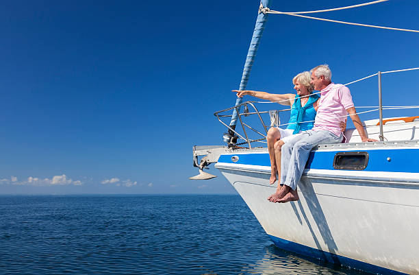 Happy Senior Couple Sailing on a Sail Boat A happy senior couple sitting on the side of a sail boat on a calm blue sea looking and pointing to a clear horizon ships bow photos stock pictures, royalty-free photos & images