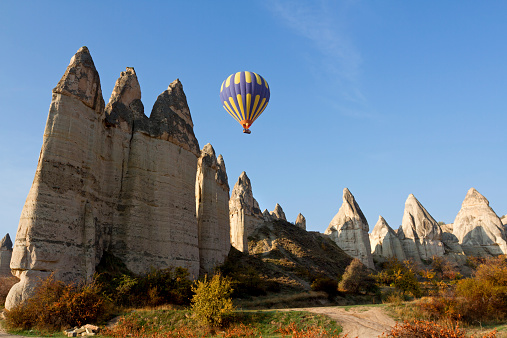 Hot air balloon carrying tourists fly over magical rocks in the Love valley in Cappadocia, Turkey. The unique shapes are a result of volcanic activity in the past. The rocks are lit by the soft morning autumn sun. The sky is clear and the landscape saturated by the autumn colors.