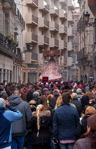 Malaga, Spain - March 21, 2016: last float in the procession of Gitanos brotherhood leaving Plaza del Teatro into a narrow street, followed by hundreds of spectators. The float with a statue of Virgin Mary is covered with plastic to protect the old and  valuable embroidery against the rain. 