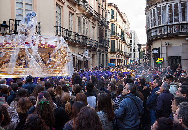 Malaga, Spain - March 21, 2016: procession of Gitanos brotherhood passing Plaza del Teatro. In spite of the bad, rainy wheather, the streets are packed with spectators looking at a float with a statue of Jezus Christ, wrapped in plastic to protect this costly statue against the rain, being carried by six rows of men dressed in purple robes and headkerchiefs. 