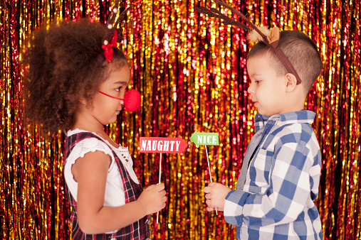 Cute little girl and boy in front of tinsel holding Christmas props on stick. They wearing funny head band with ears of reindeer and holding arrows facing each other with the inscription naughty and nice.