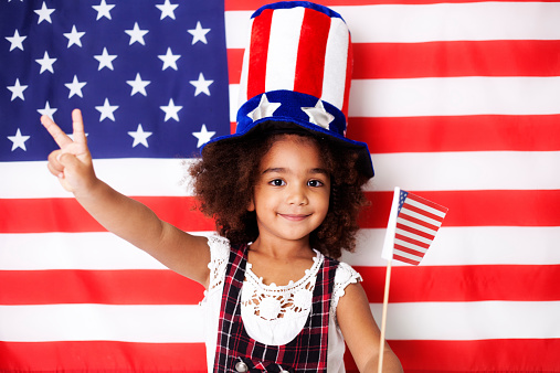 Pretty little girl with patriotic hat in front of American flag gesturing victory and holding USA flag.
