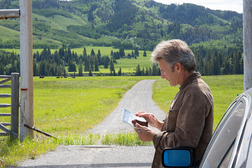 Man relaxes besides car, uses digital tablet, ranchlands