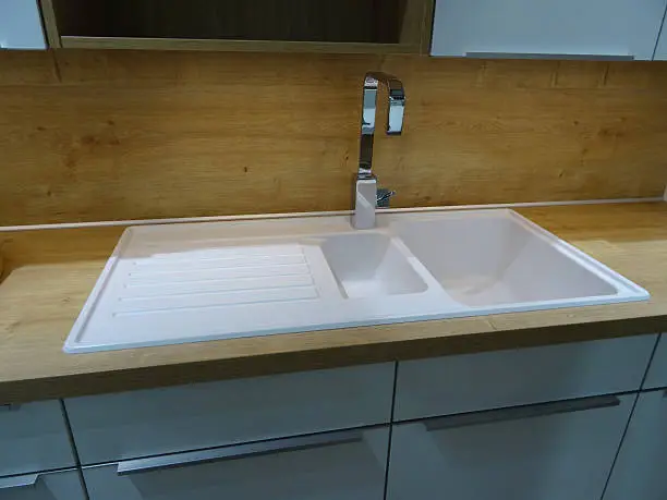 Photo showing a modern double white ceramic kitchen sink and drainer / draining board with a brushed stainless-steel mixer tap.  The sink is part of a contemporary kitchen with a wood effect laminate worktop counter (oak countertop), and is positioned beneath some duck egg grey glossy wall cabinets, with brushed chrome handles and shiny doors.  Under cupboard strip lighting and a solid wooden / oak wood splashback panel (rather than kitchen tiling) complete this domestic scene.