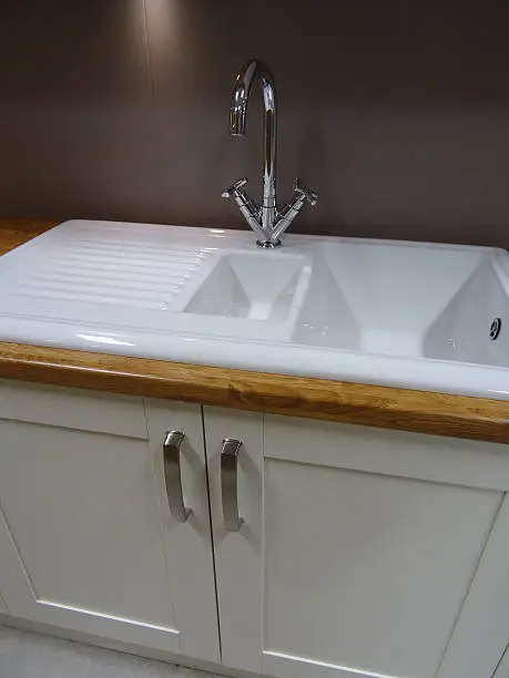 Photo showing a modern double white ceramic kitchen sink and drainer / draining board with a brushed stainless-steel mixer tap.  The sink is part of a contemporary kitchen with a wood effect laminate worktop counter (oak countertop), and is positioned beneath some duck egg grey wall cabinets, with brushed chrome handles.  Under cupboard strip lighting and a solid splashback panel (rather than kitchen tiling) complete this domestic scene.