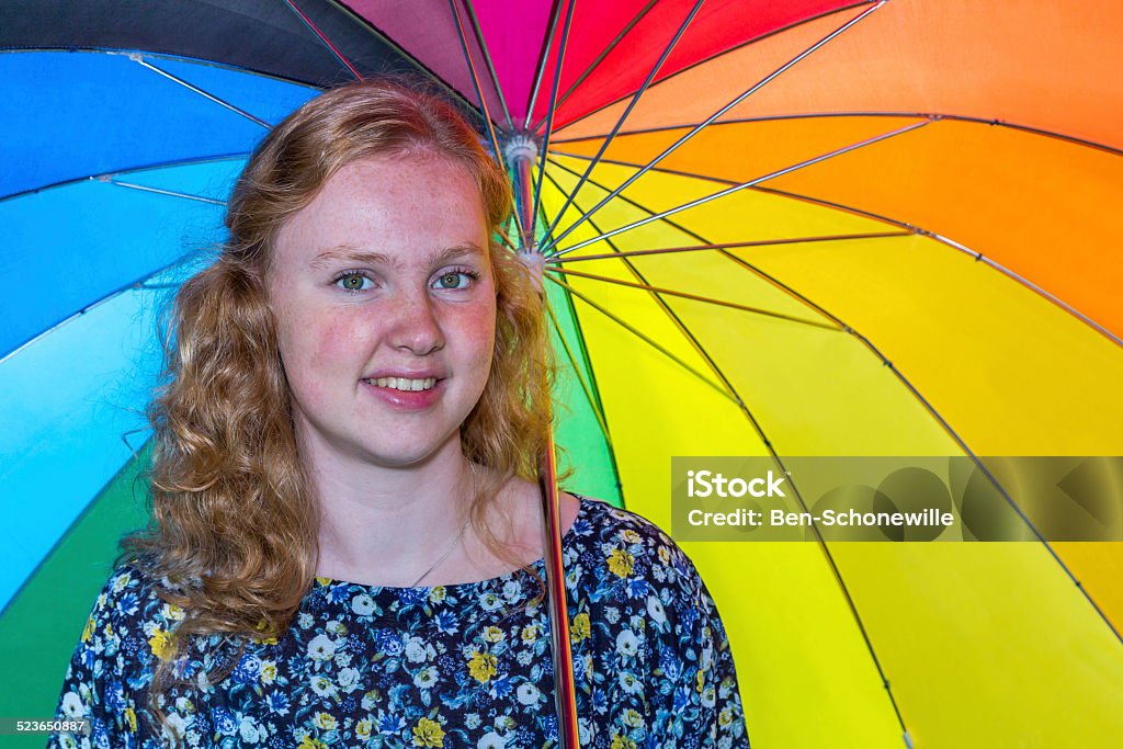 Teenage girl under colored umbrella Dutch teenage girl under umbrella with various different colors like rainbow. Caucasian girl with long hair posing and smiling at the camera. She holds the colorful umbrella behind her. Bad weather like rain will not affect her. The background is bright and outstanding Adolescence Stock Photo