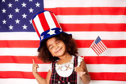 Pretty little girl , have closed eyes,  with patriotic hat in front of American flag holding USA flag and showing with finger up