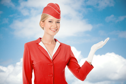 Charming Stewardess Dressed In Red Uniform Holding In Hand. Sky With Clouds Background.